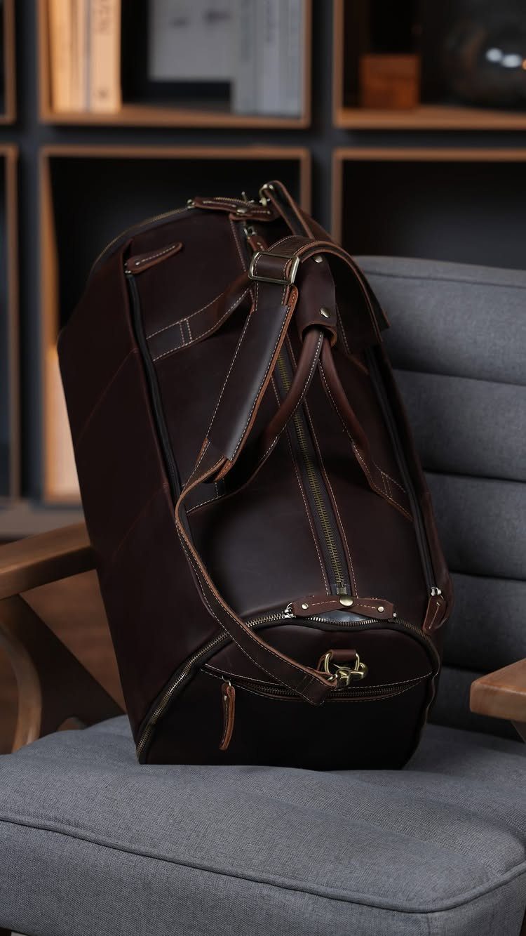 Dark brown leather duffel bag sitting on a grey fabric armchair