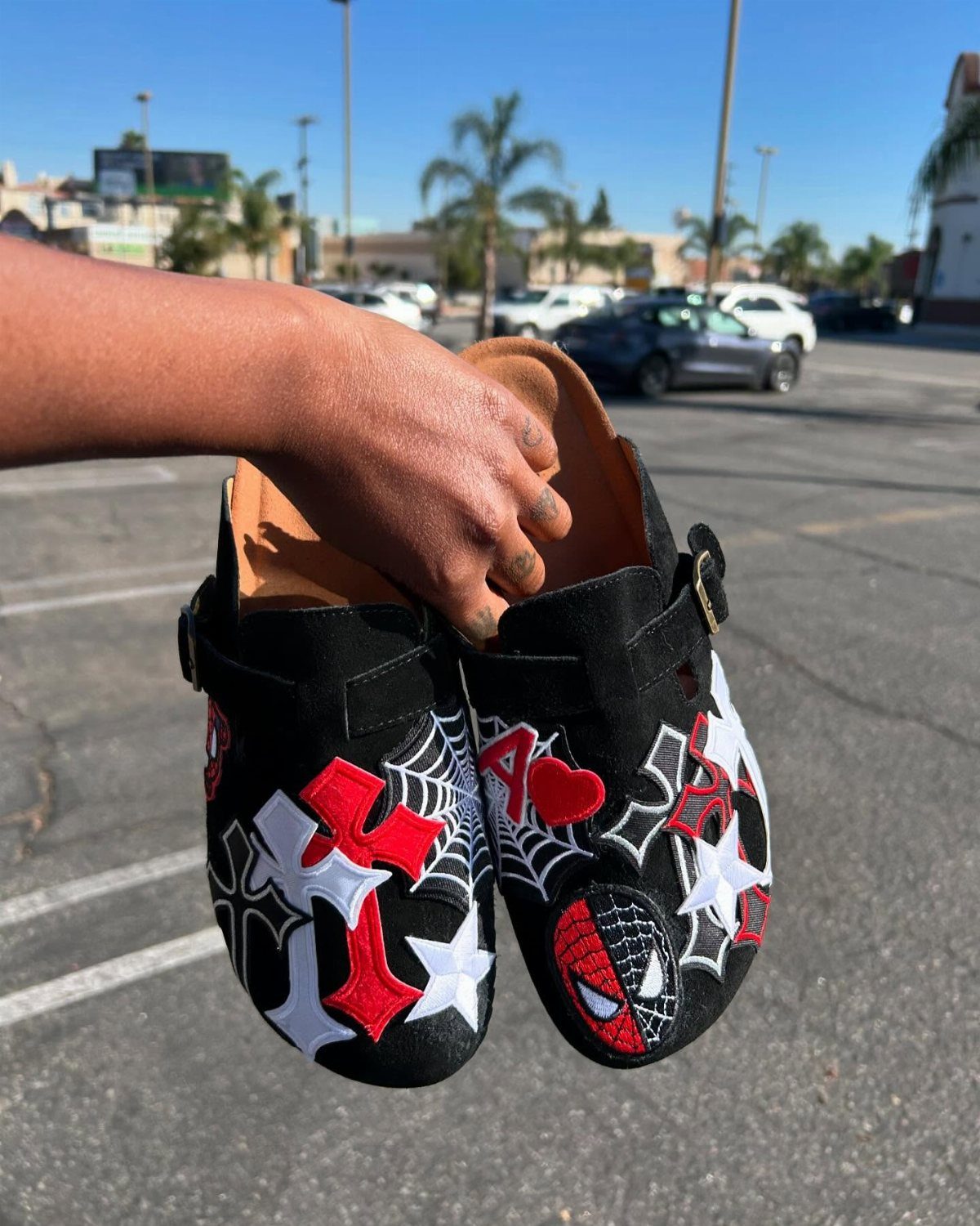 Black suede clogs with red and white Spider-Man embroidery held in a hand.