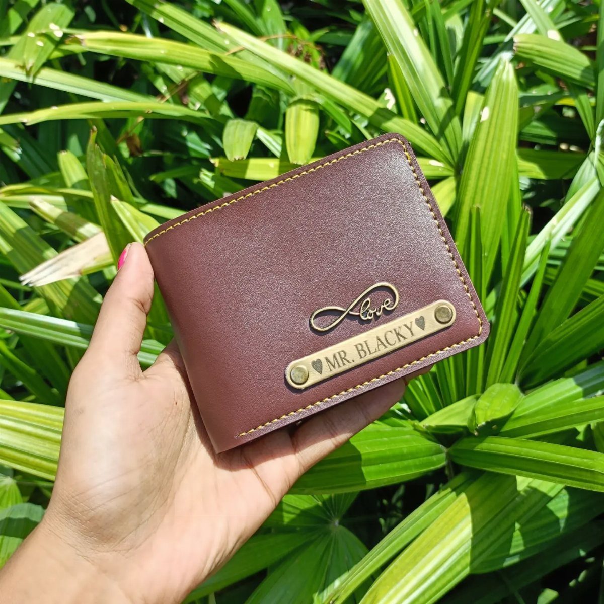 Brown leather bifold wallet with personalized gold metal plate held against green leaves.