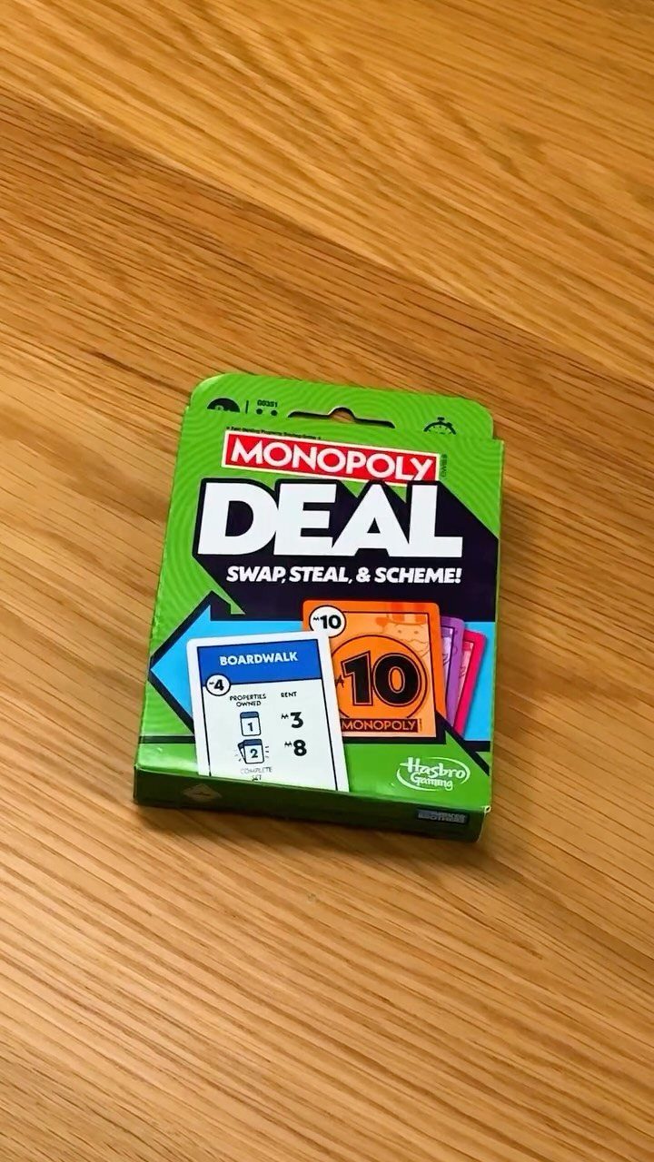 Monopoly Deal card game box sitting on a light brown wooden table surface.