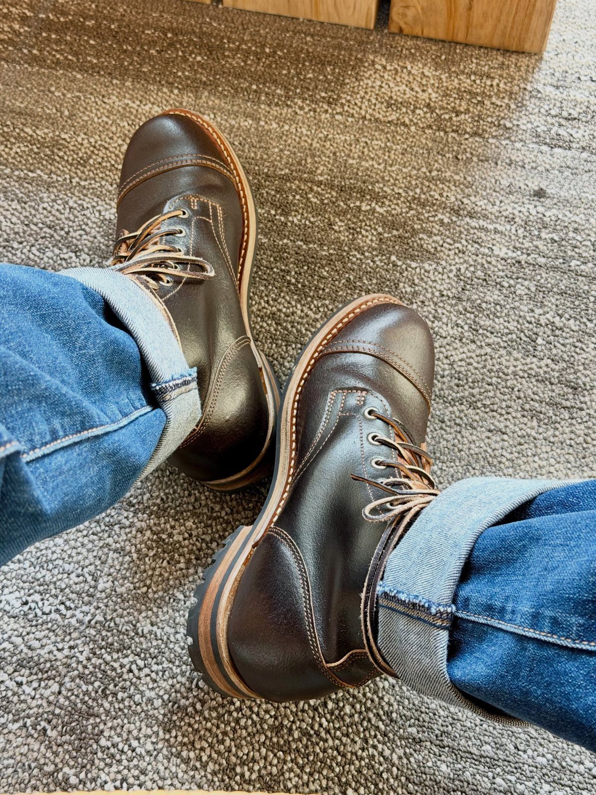 Dark brown waxed leather boots worn with cuffed blue jeans on grey carpet.