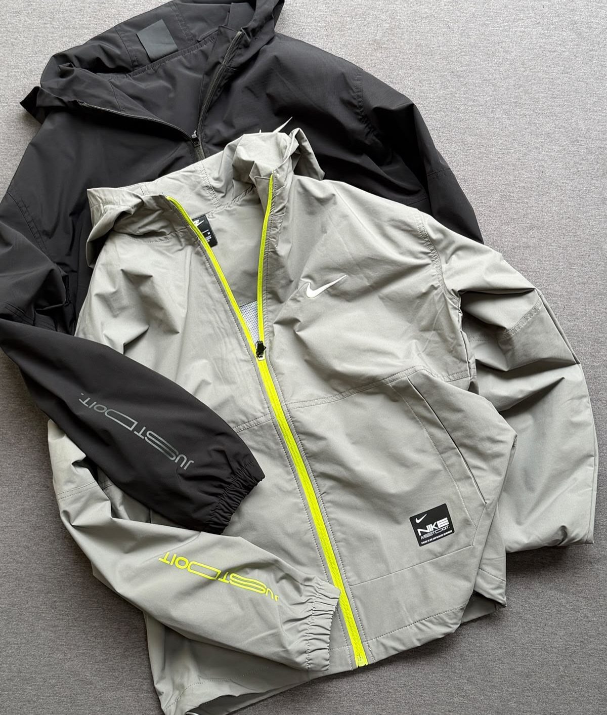 Two athletic windbreaker jackets, one black and one grey with yellow zipper, flat lay.