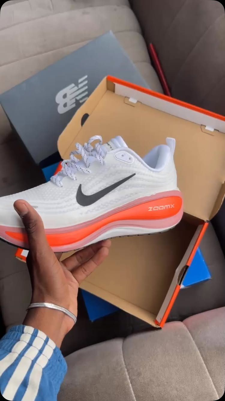 White Nike running shoe with orange sole held over an open shoe box.