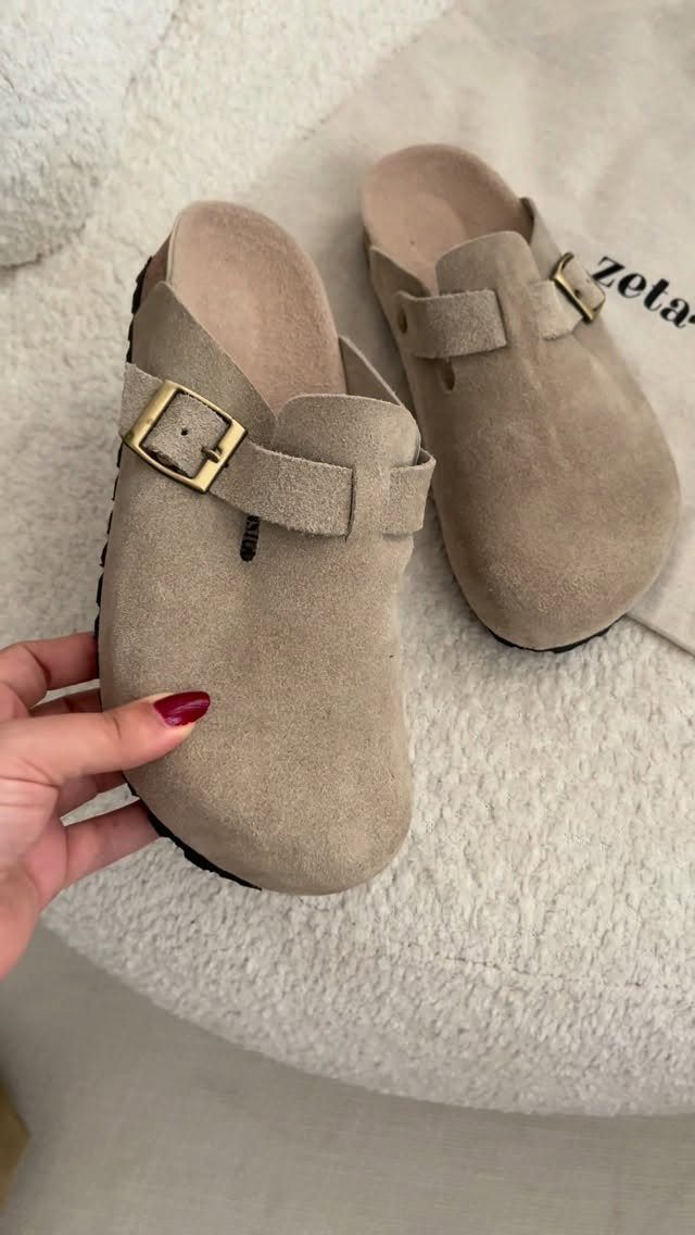 Beige suede clog shoes with gold buckles held over a white fluffy rug.