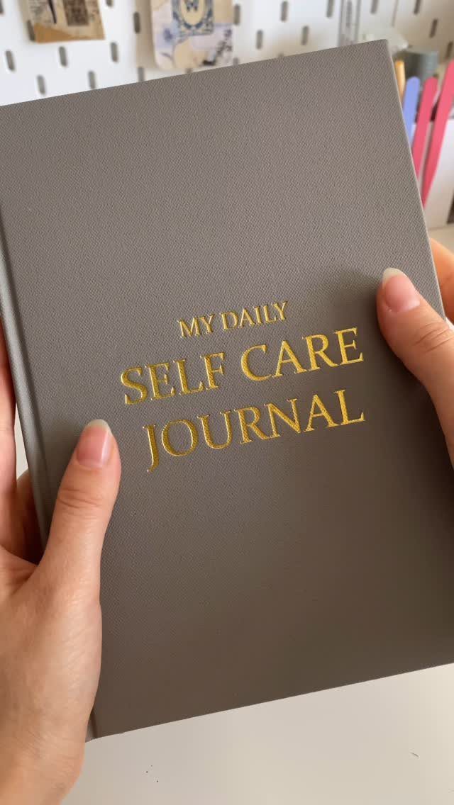Grey self care journal with gold lettering held by two hands on white desk
