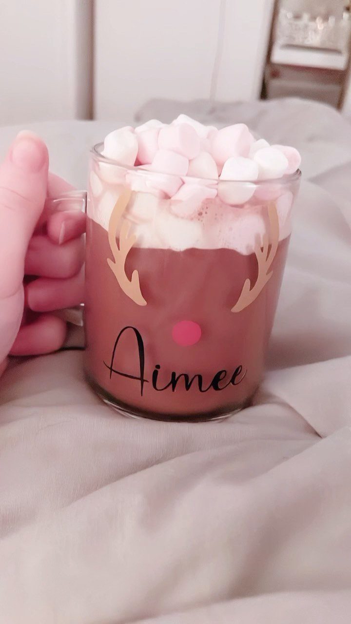 Glass mug of hot chocolate with marshmallows and reindeer design on beige fabric.