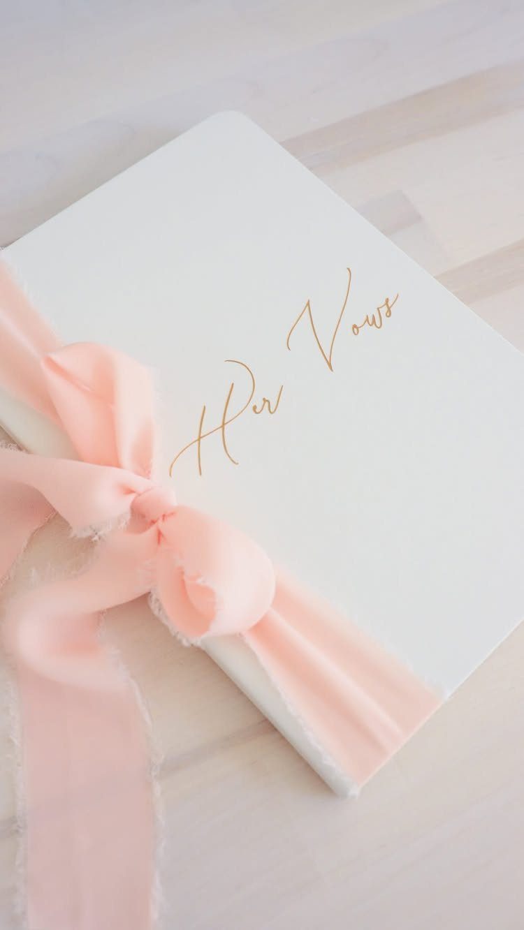 White vow book with gold script and a pink silk ribbon bow.