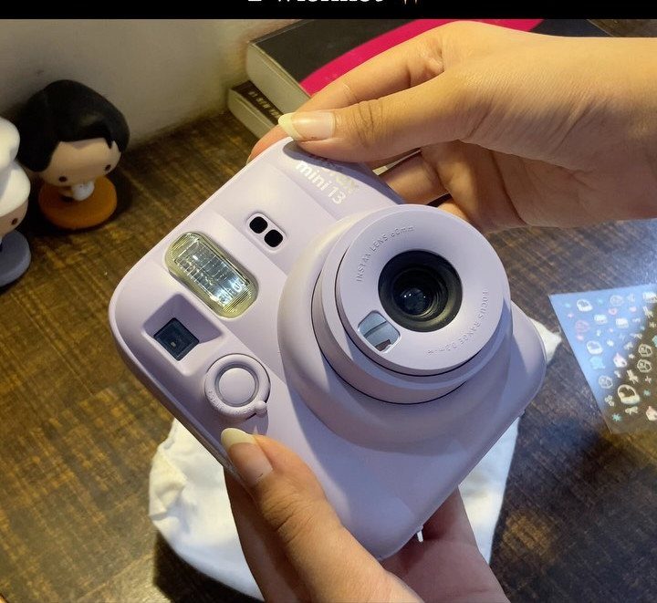 Light purple instant camera held by two hands over a wooden table surface.