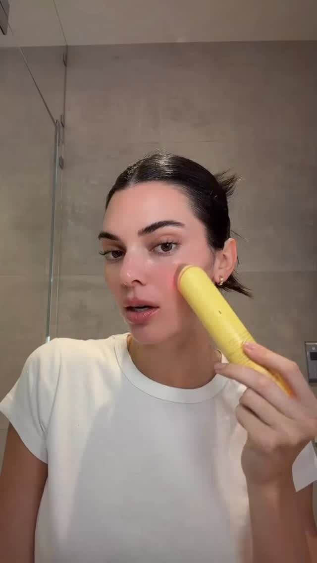 Woman using a yellow depuffing wand on her cheek in a bathroom.