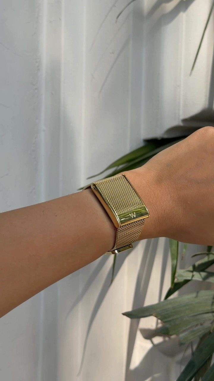 Gold mesh metal watch on a wrist against a white wall and greenery
