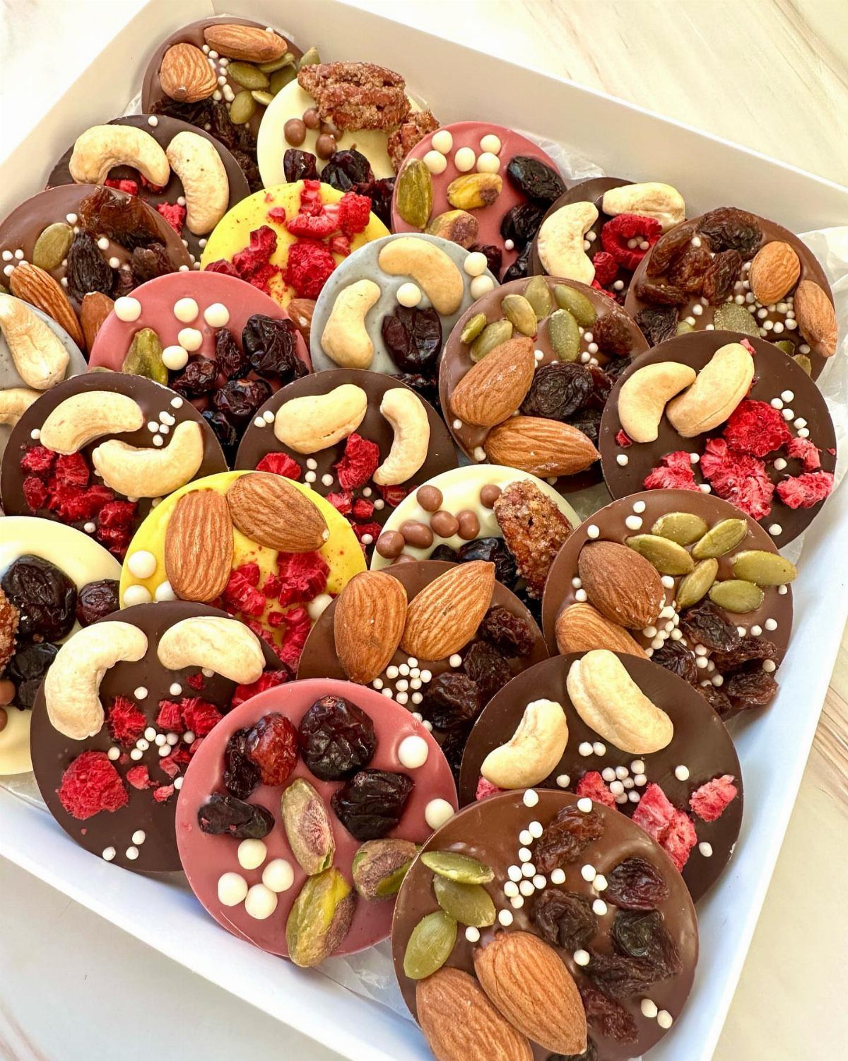 Assorted round chocolate discs topped with nuts and dried fruits in a white box.