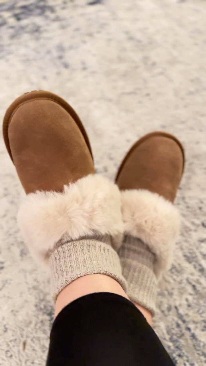 Brown suede slippers with white faux-fur trim and grey socks on a rug.