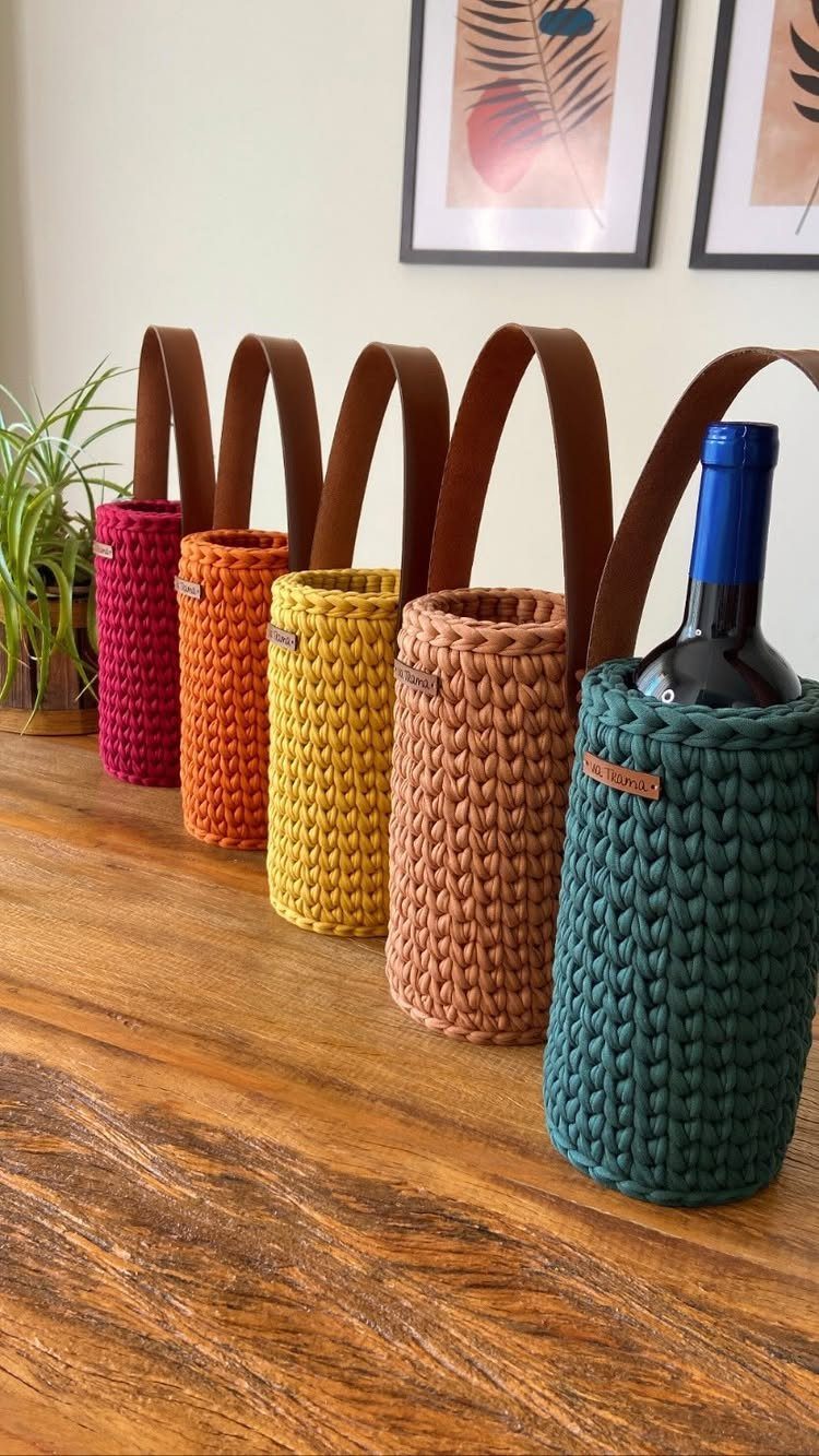 Knitted wine bottle carriers in various colors with leather handles on a wooden table