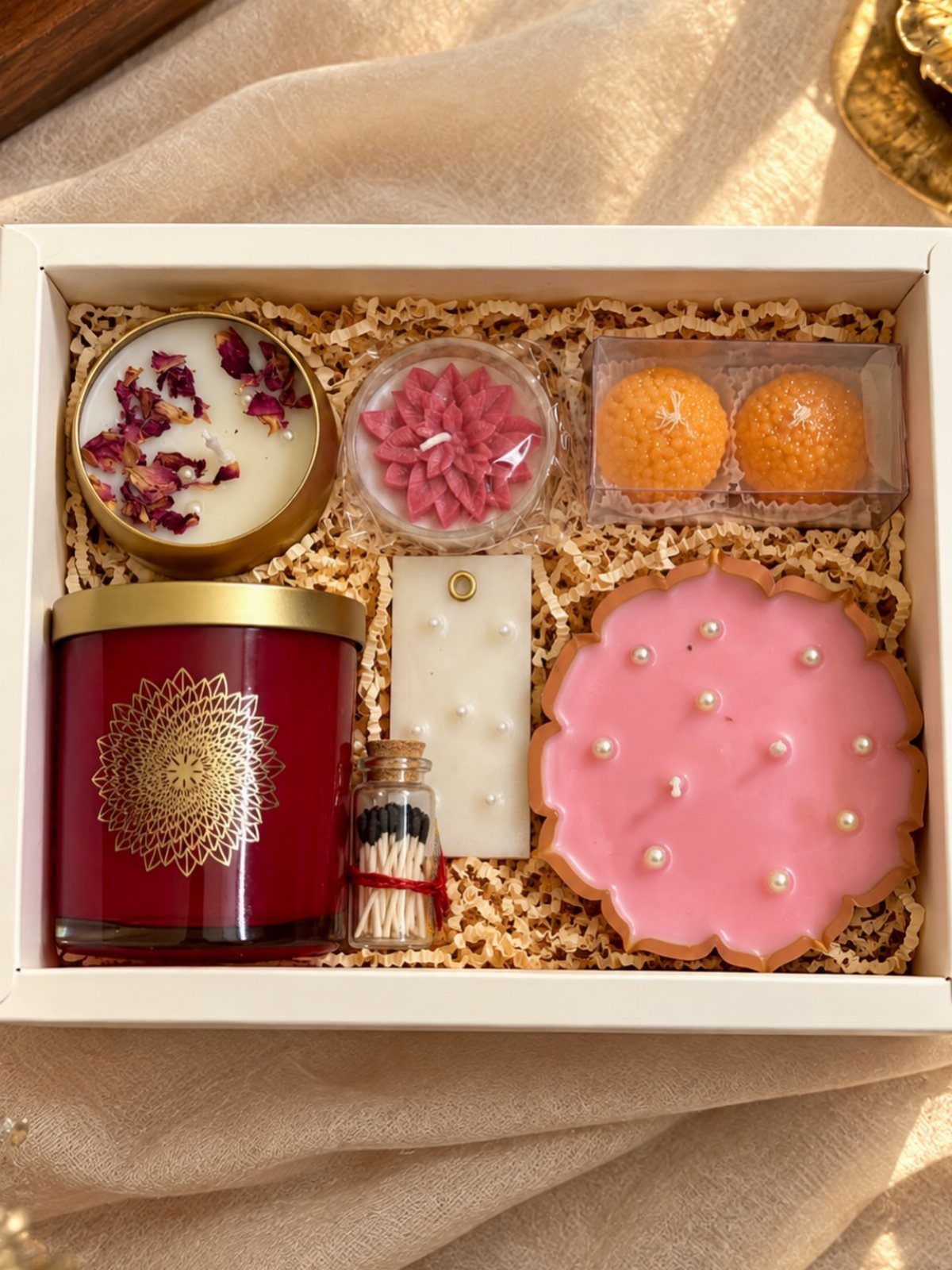 Gift box containing various decorative candles, matches, and treats on shredded paper.