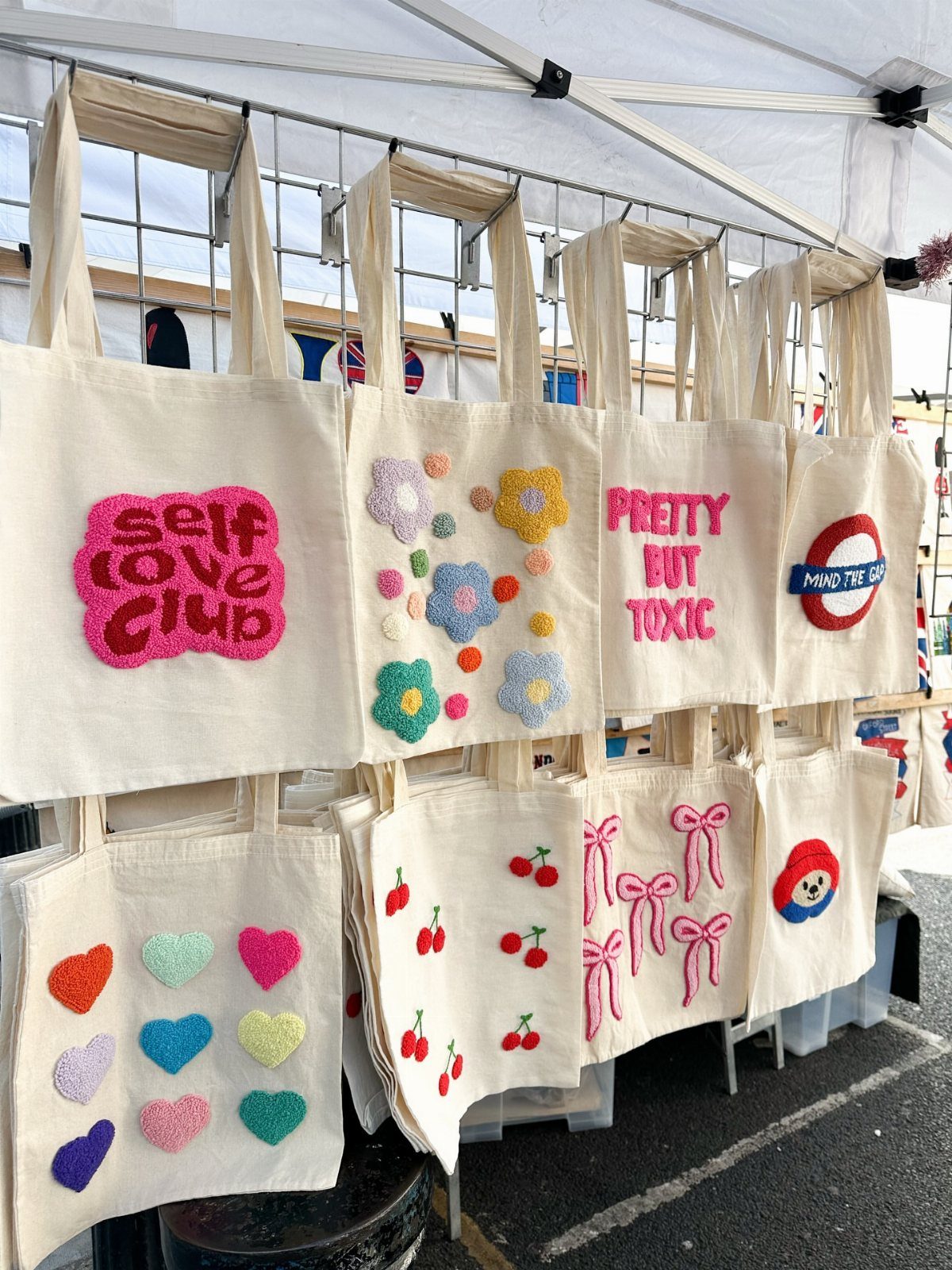 Cream canvas tote bags with colorful tufted designs hanging at an outdoor market.