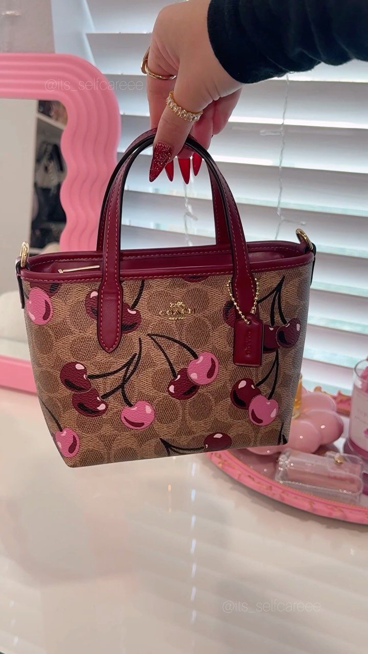 Brown Coach handbag with cherry print held by a hand with red nails.