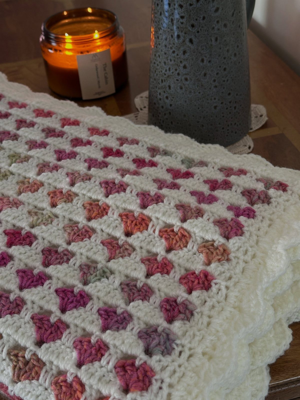 White crocheted blanket with colorful heart patterns next to a lit candle.