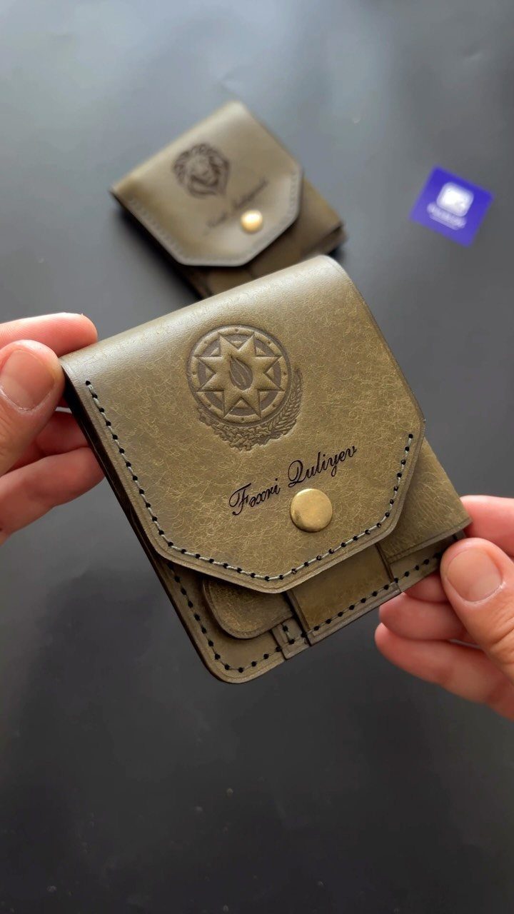 Olive-green leather wallet with an engraved name and circular emblem held by hands.