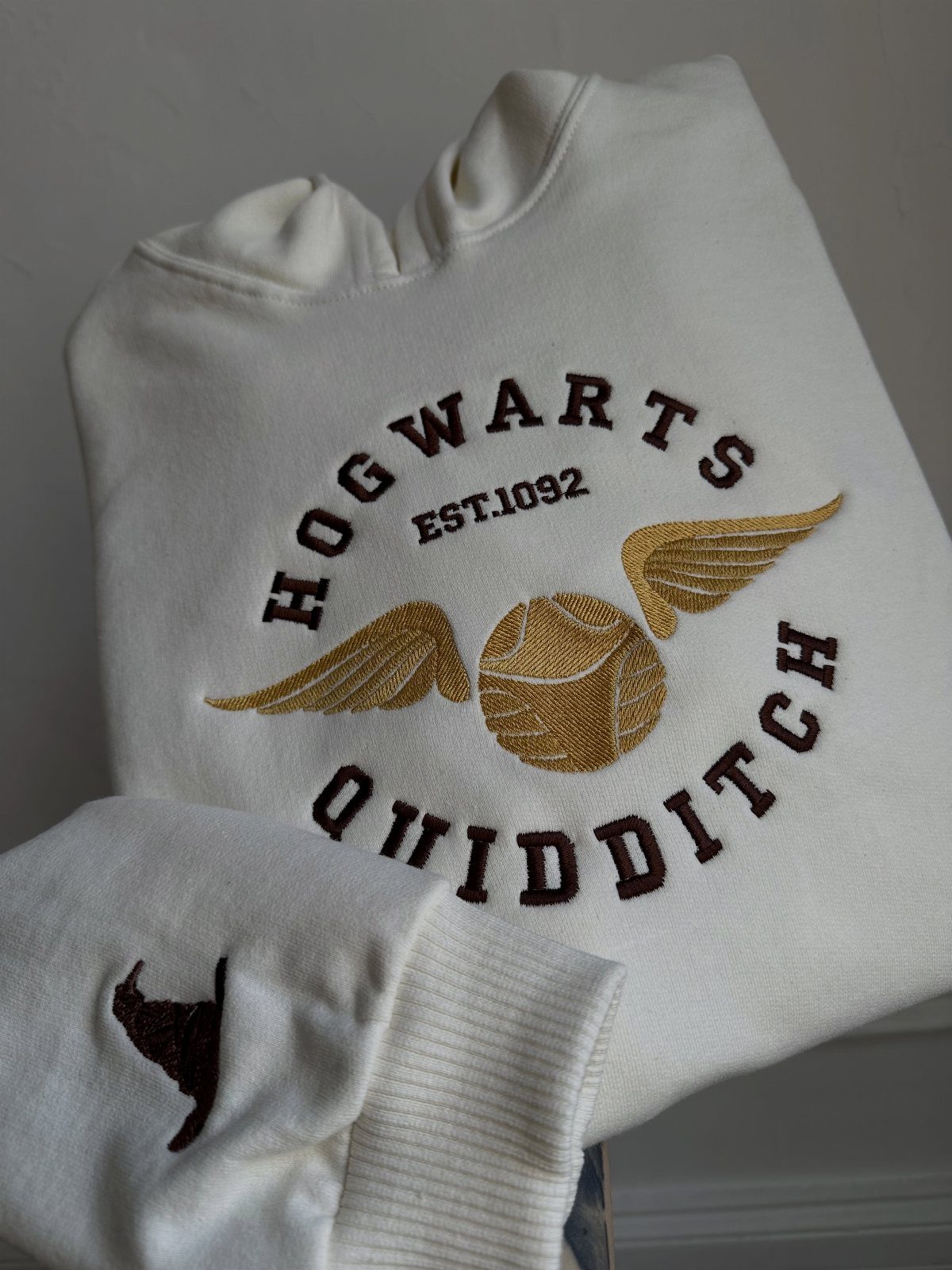 25 Best Harry Potter Gifts for Every Wizarding Fan in 2026 2 White Hogwarts Quidditch hoodie with a golden snitch embroidery and brown text.