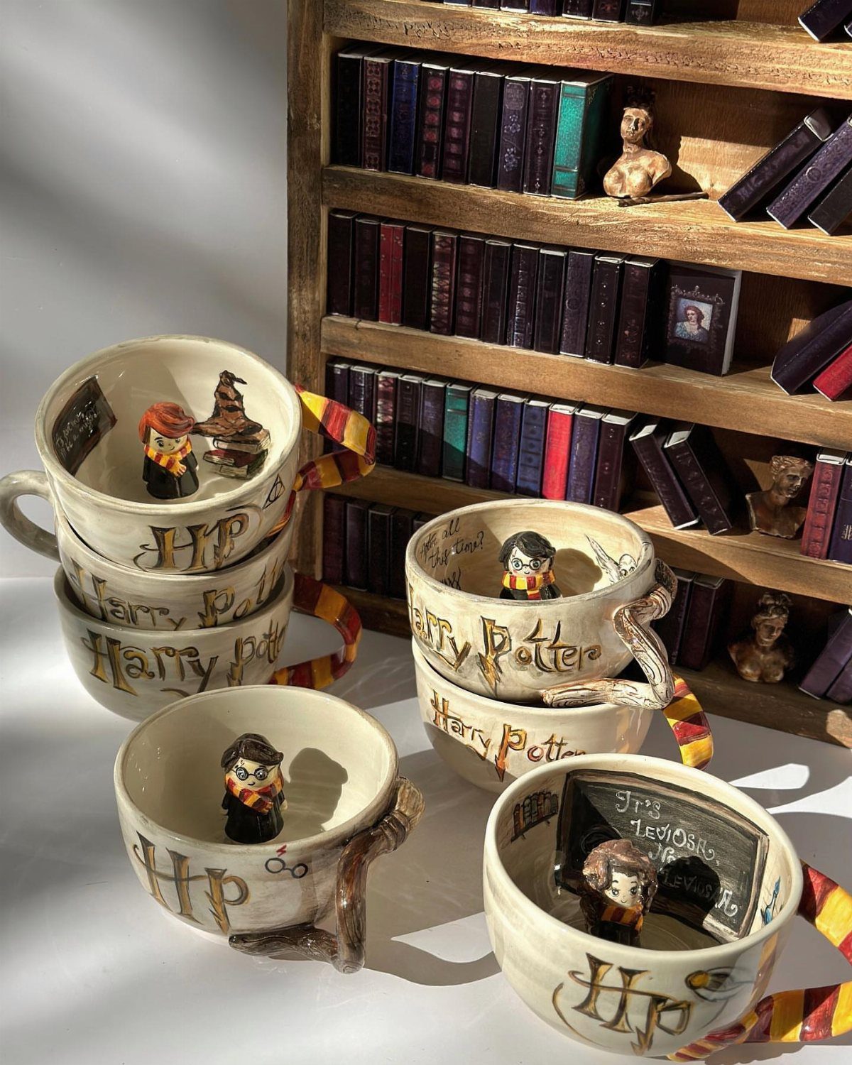 25 Best Harry Potter Gifts for Every Wizarding Fan in 2026 3 White Harry Potter mugs with character figurines and a miniature bookshelf background.
