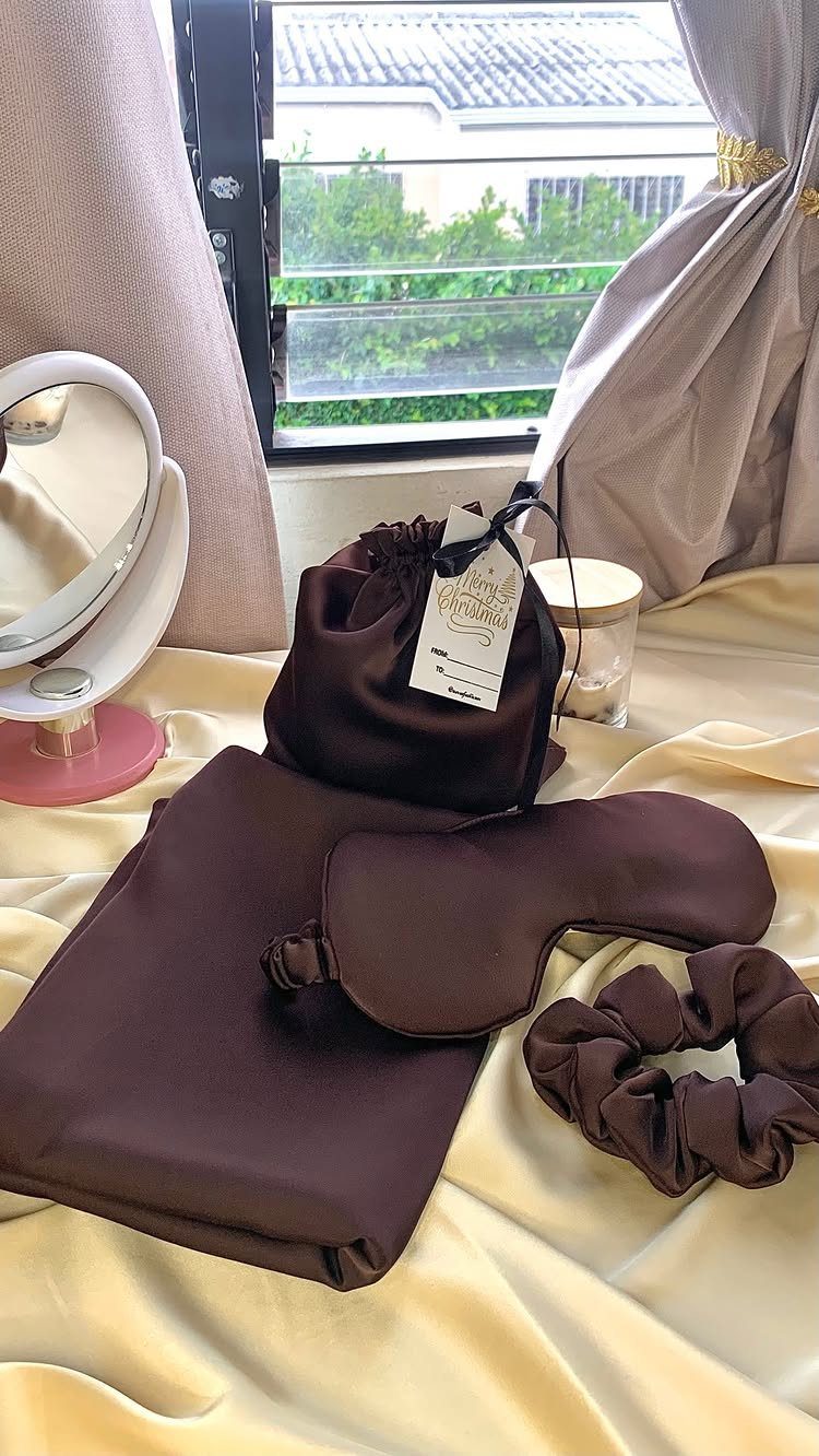 Brown satin sleep set with pillowcase, eye mask, and scrunchie on cream fabric.