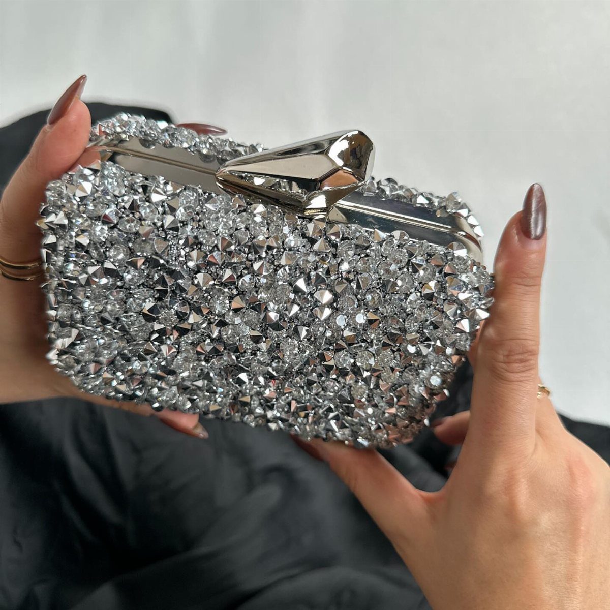 Silver crystal-covered clutch bag held by hands with metallic manicured nails.