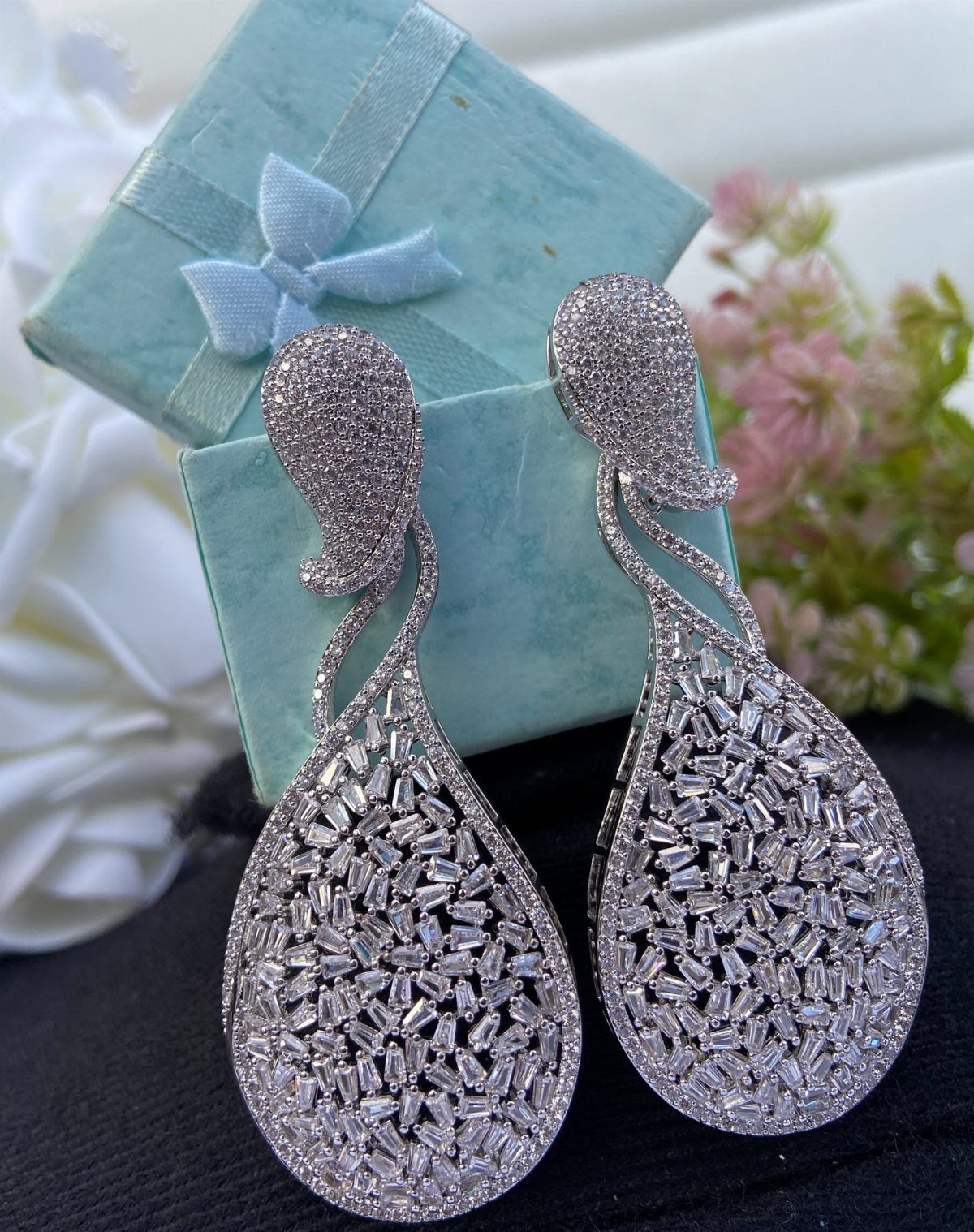 Large teardrop silver earrings with crystals next to a blue gift box and flowers