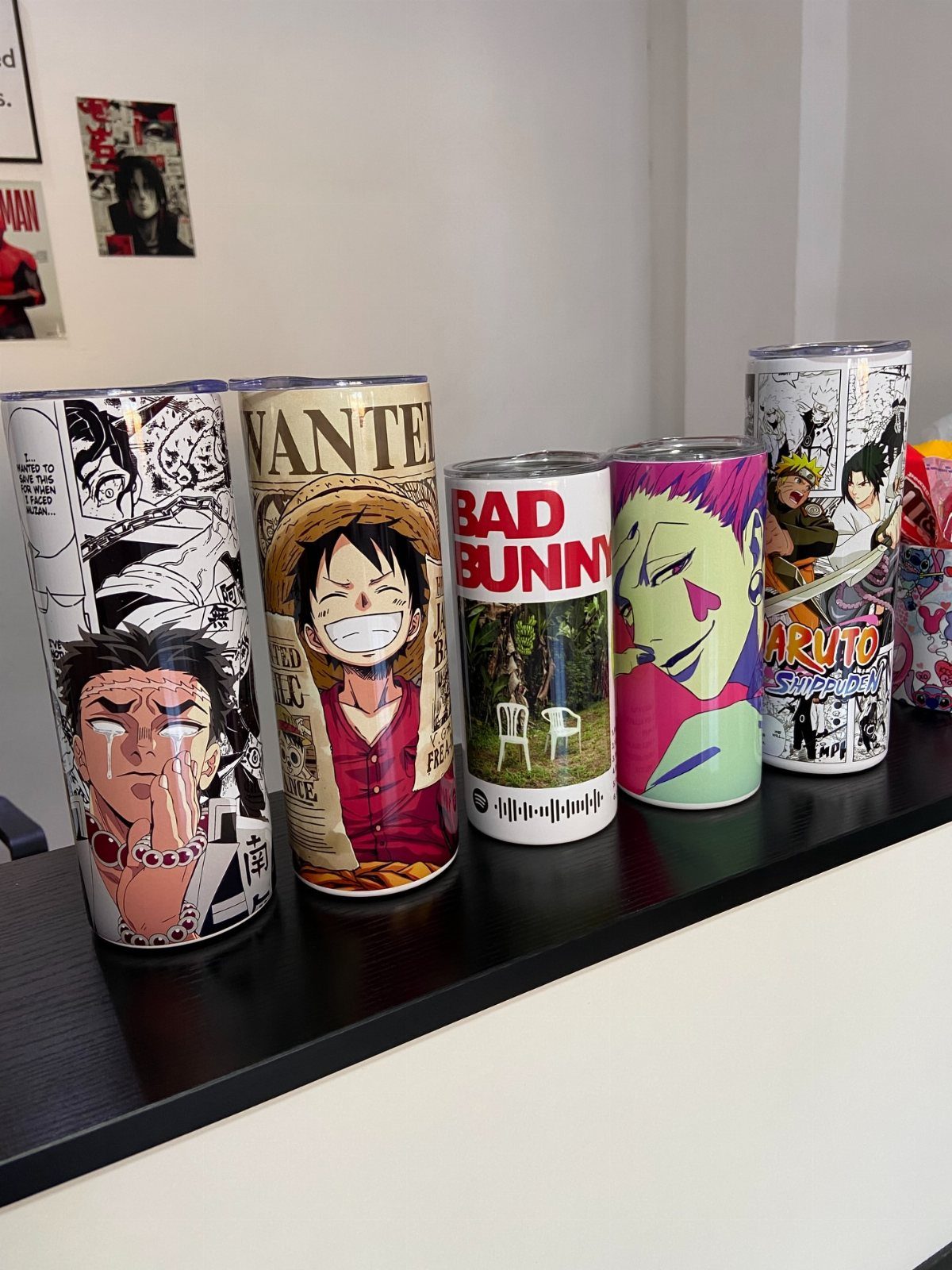 Five customized travel tumblers with anime and music graphics on a black shelf.