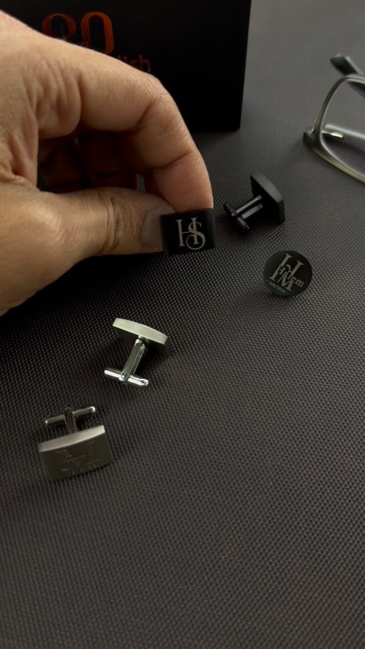 Hand holding a black engraved cufflink among other personalized cufflinks on grey surface