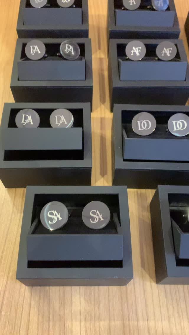 Silver monogrammed cufflinks displayed in black boxes on a wooden table.