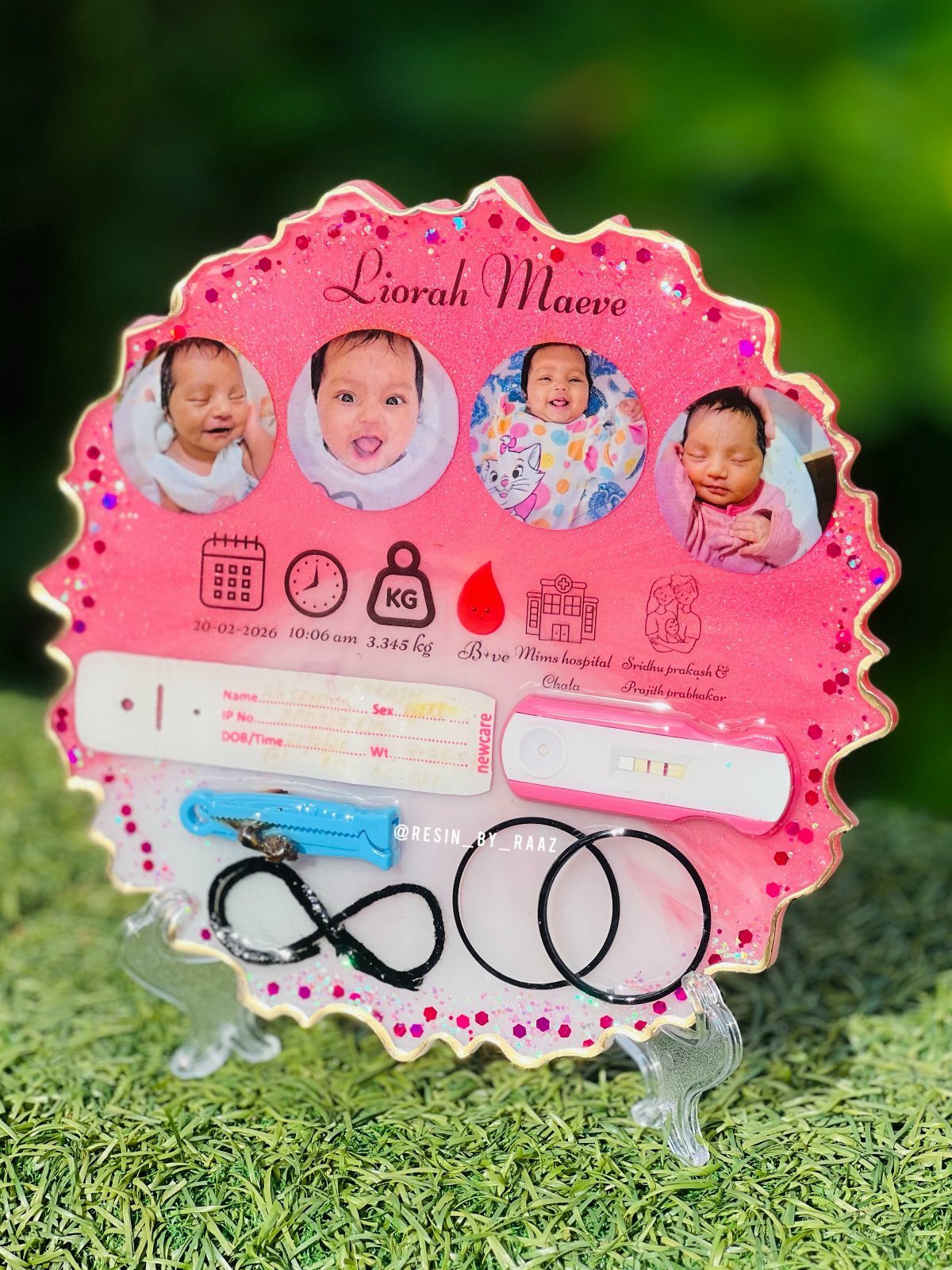 Pink glittery resin baby keepsake plaque with photos and birth memorabilia on grass.