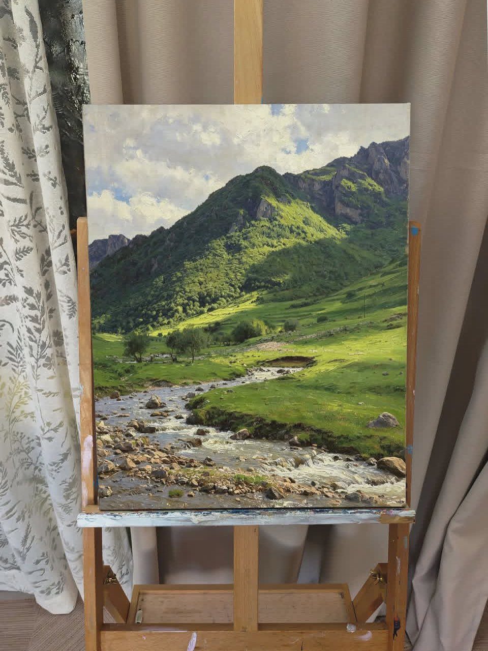 12 Paintings as Gifts for Every Budget and Taste 3 Landscape painting of green mountains and a river on a wooden easel