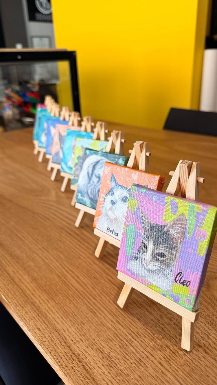 12 Paintings as Gifts for Every Budget and Taste 9 Small cat paintings on miniature wooden easels lined up on a wooden table.