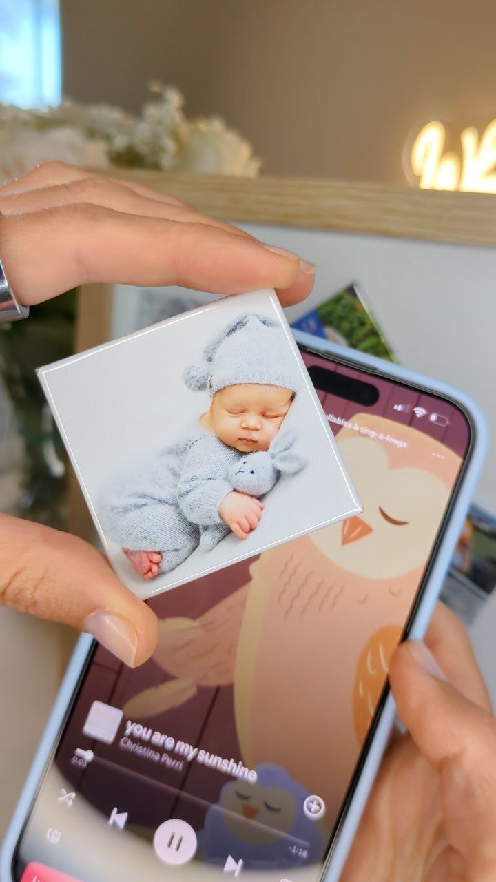 Square photo magnet of a sleeping baby held over a smartphone screen.