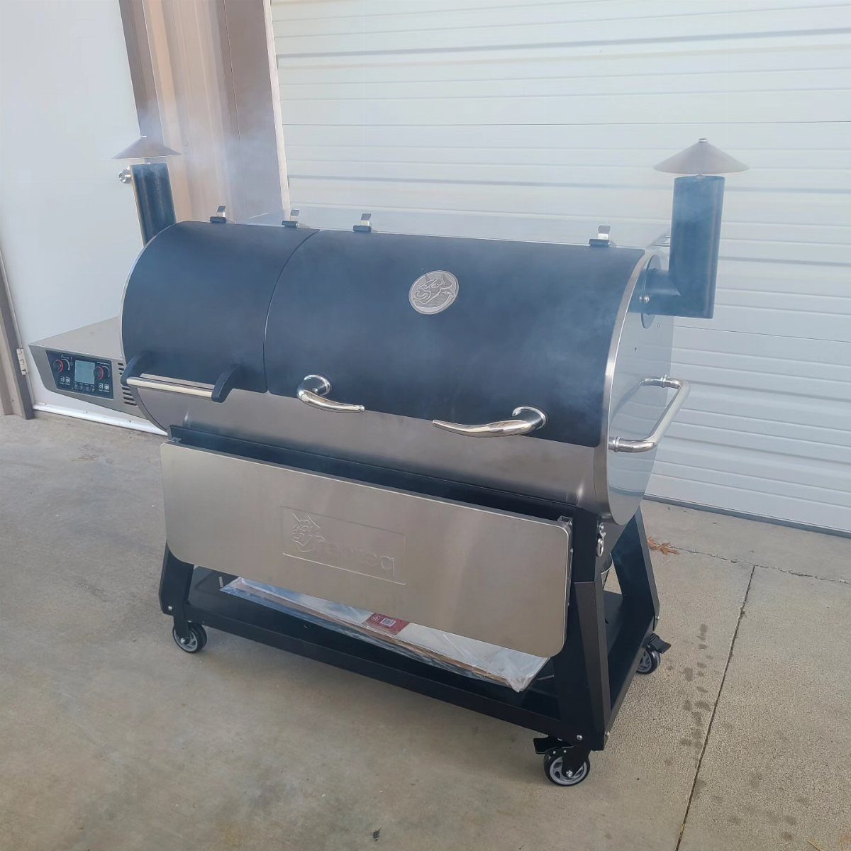Black and stainless steel pellet grill smoker emitting smoke on a concrete driveway