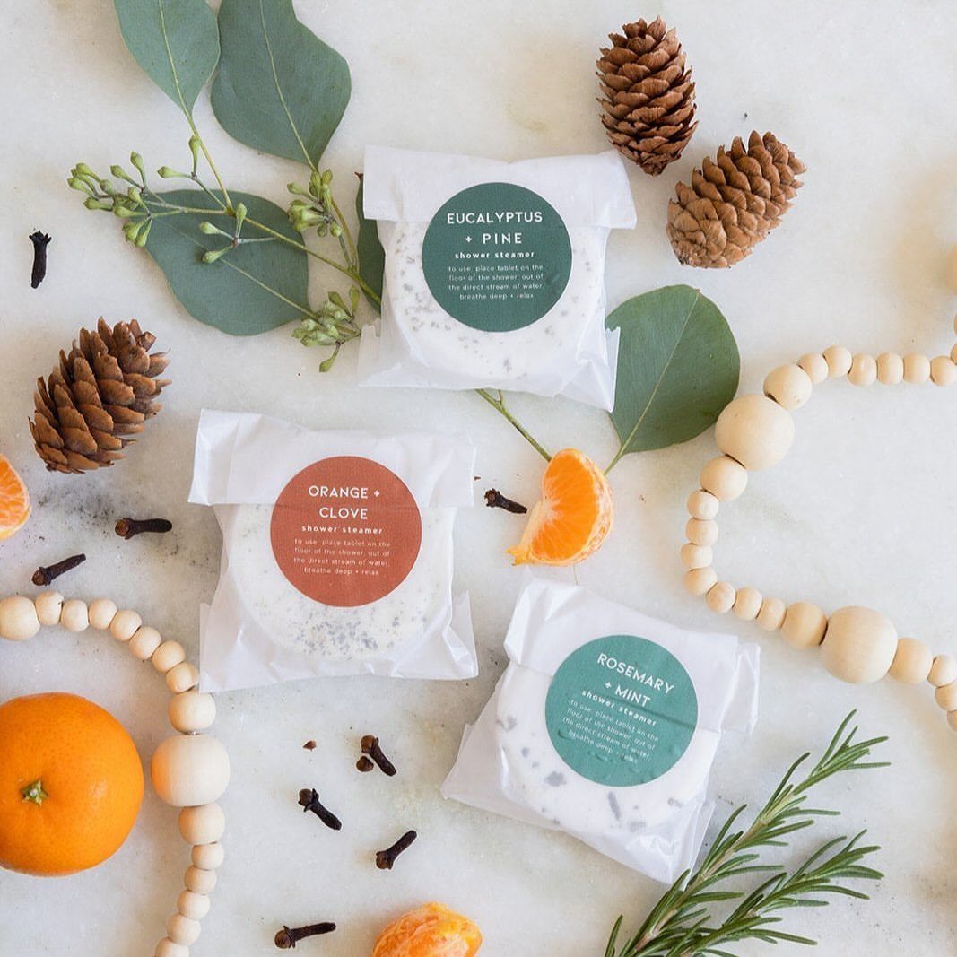 Shower steamer packets surrounded by pinecones, eucalyptus leaves, and orange slices on white.
