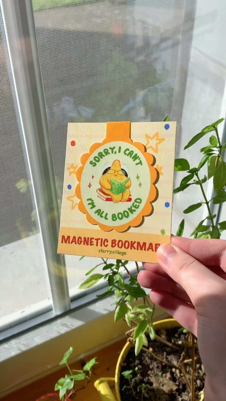 Magnetic bookmark with a reading cat illustration held against a window background.