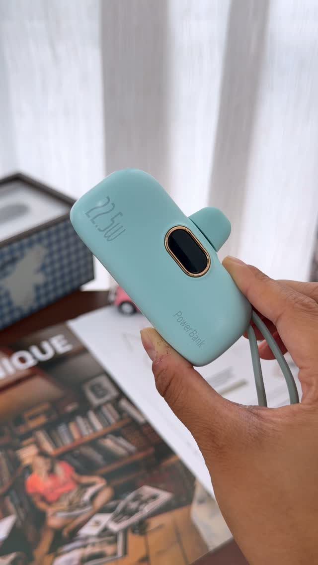 Light blue portable power bank held in a hand over a desk setting.
