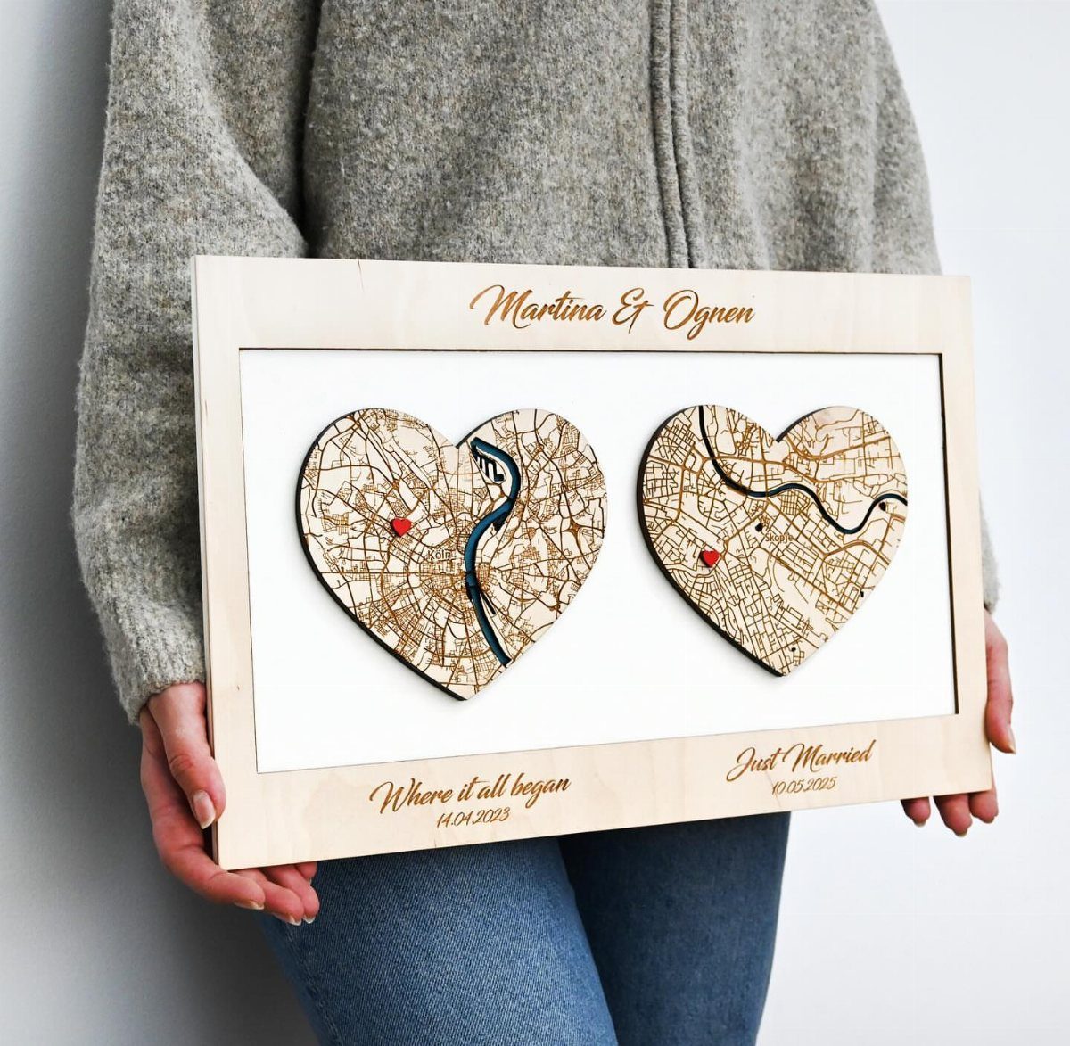 Wooden framed art featuring two heart-shaped city map engravings held by a person.