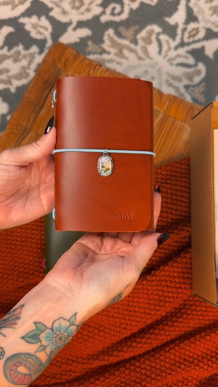 Brown leather journal with embossed initials and a blue closure band held by hands.