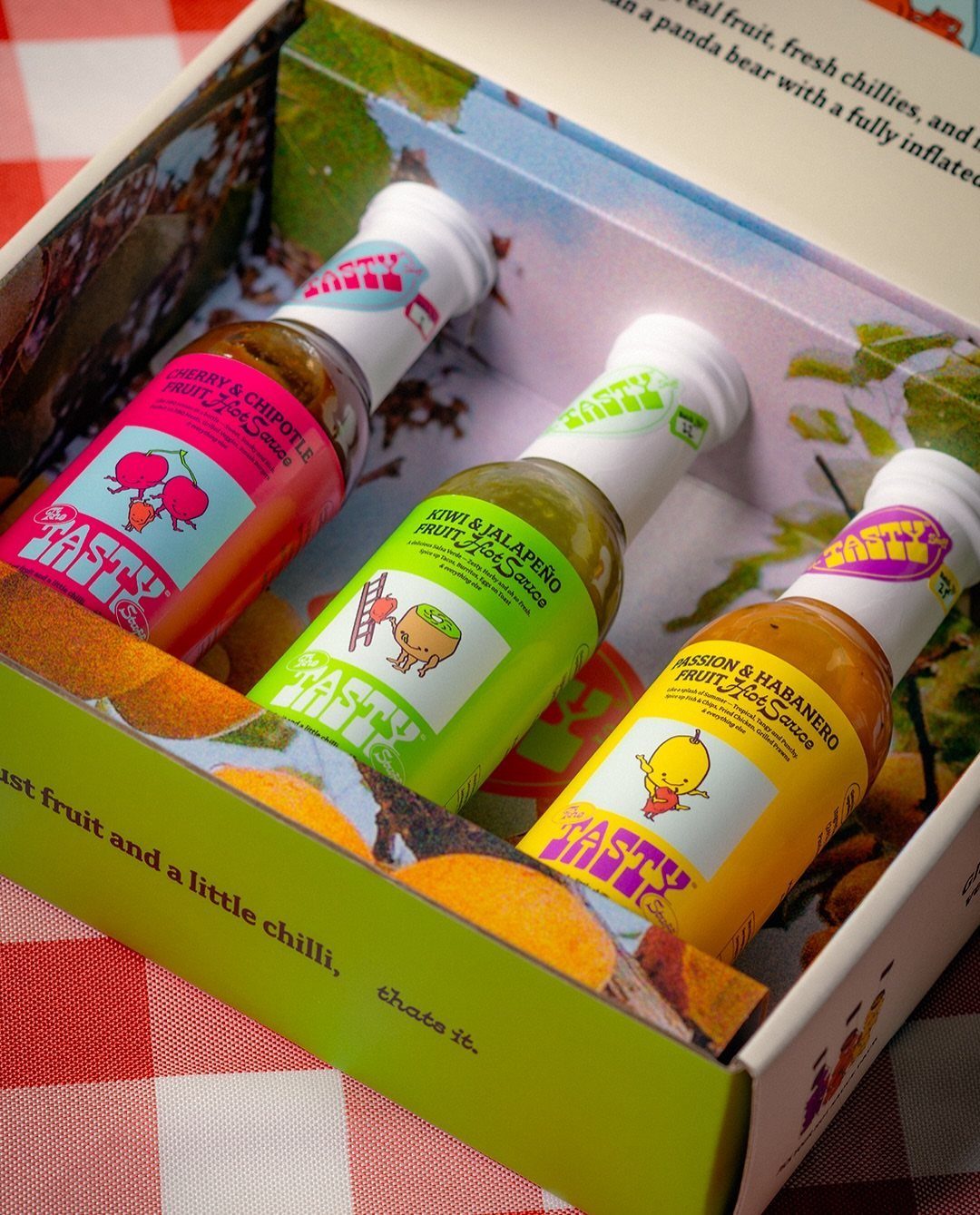 Three colorful hot sauce bottles in a decorative gift box on checkered cloth.