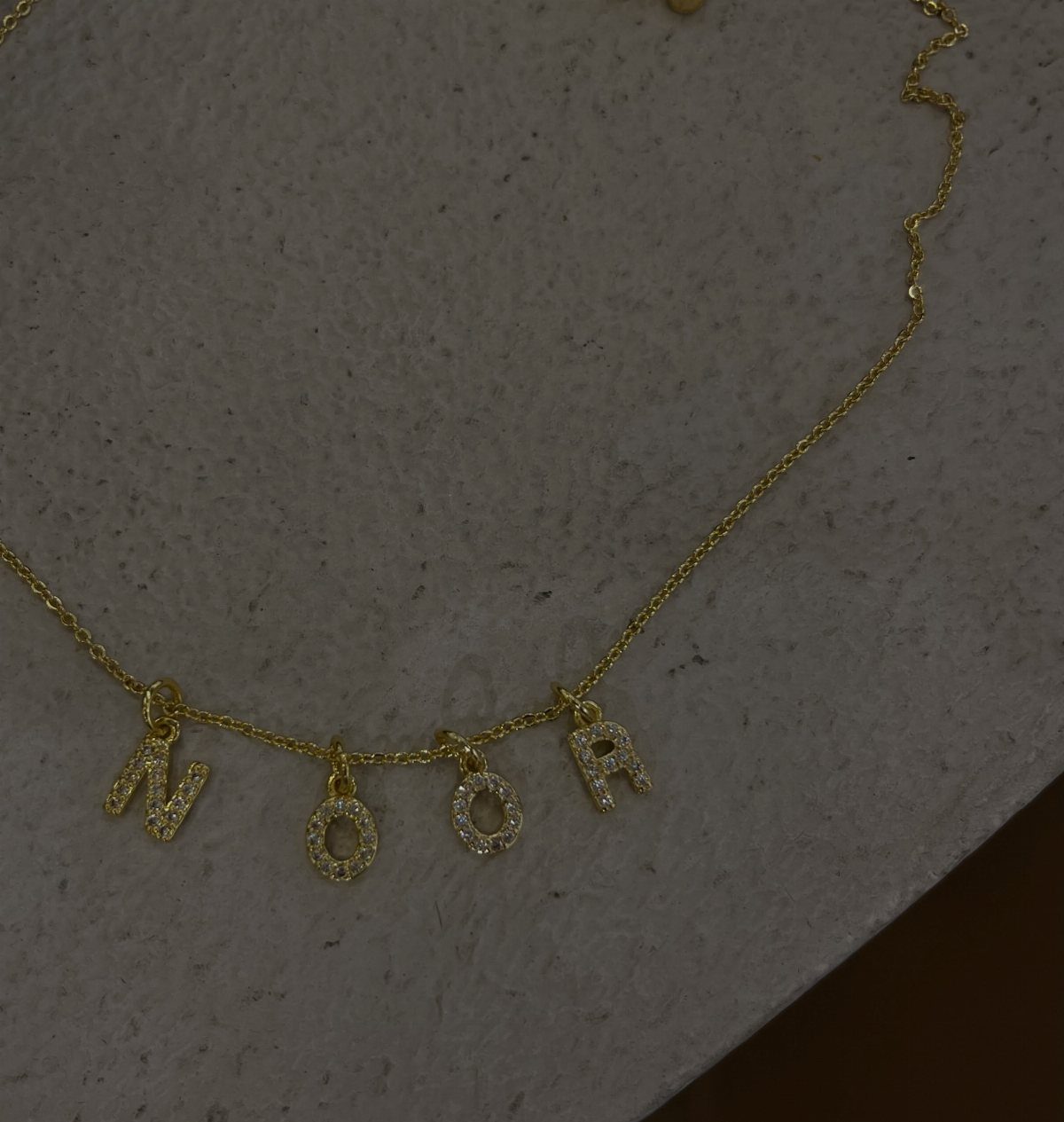 Gold necklace with sparkling letter charms spelling NOOR on a grey stone background