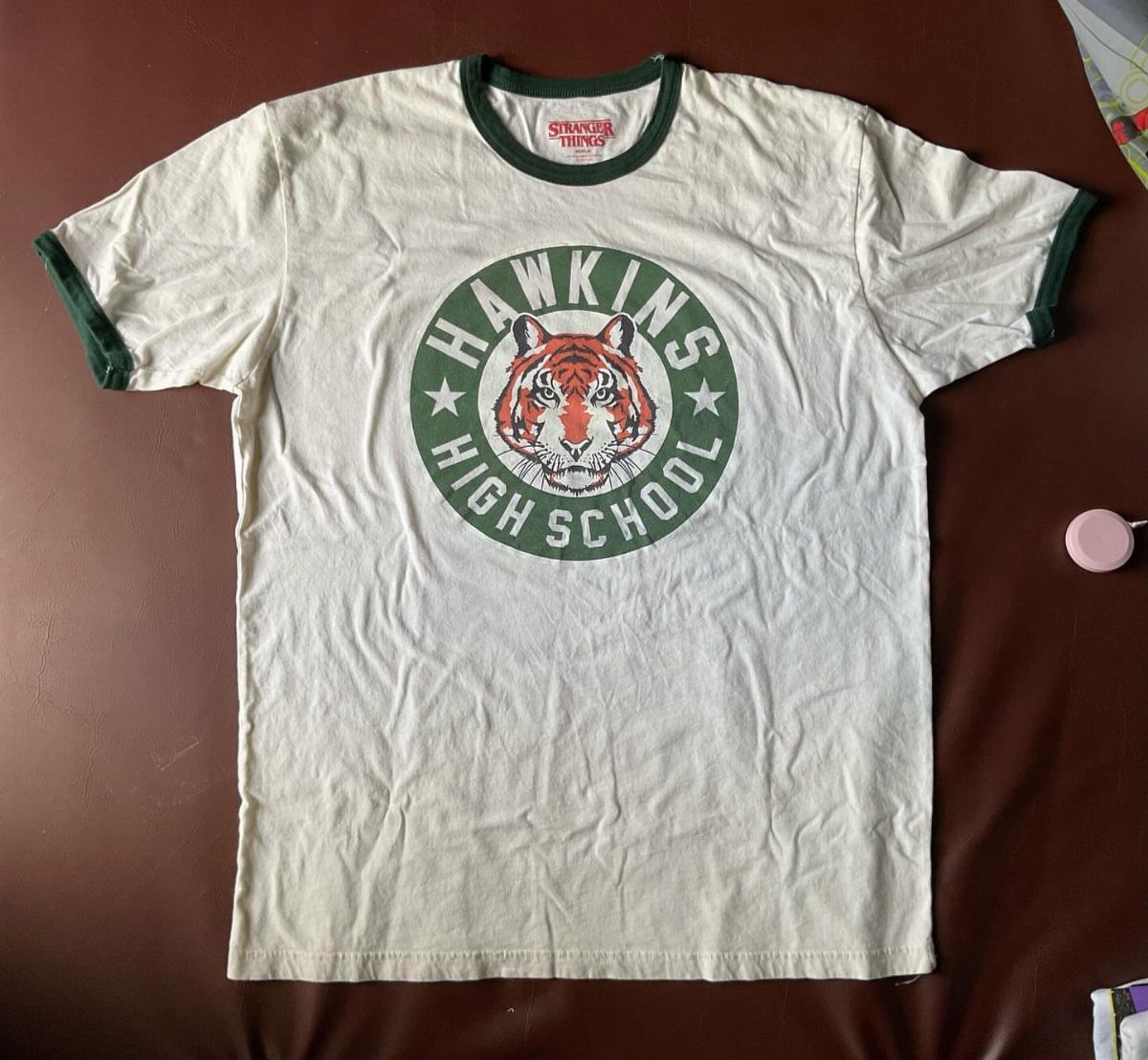 Cream t-shirt with green trim and Hawkins High School tiger logo on chest.