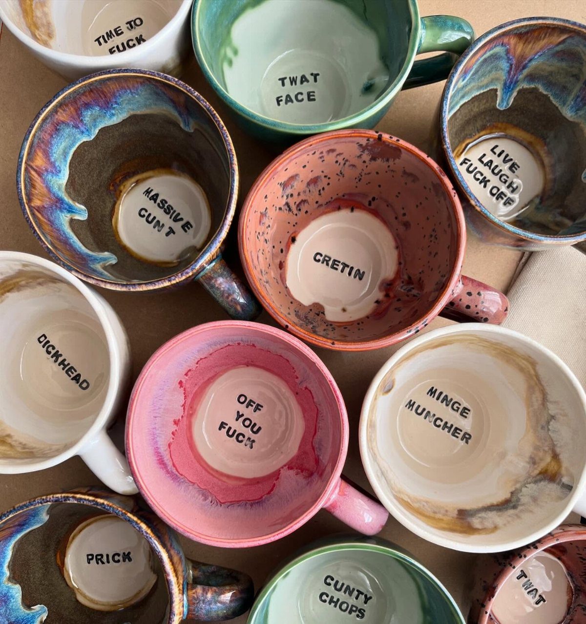 Colorful ceramic mugs with profane phrases printed on the inside bottom surfaces.