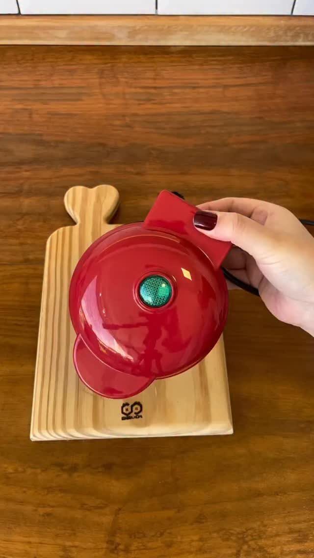 Red mini waffle maker resting on a wooden cutting board held by a hand.