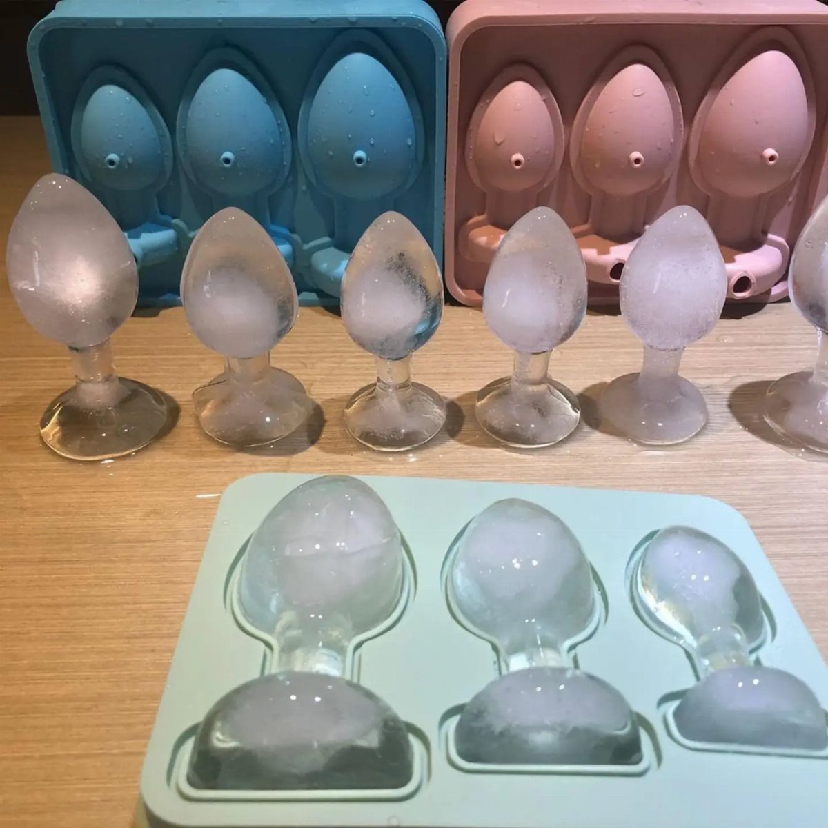 Penis-shaped ice cubes and silicone molds on a light wooden table.