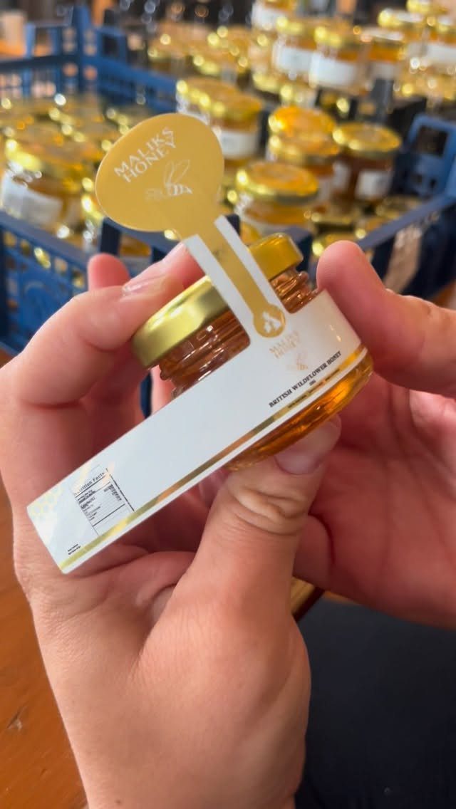 Small glass jar of golden honey held by hands with many jars behind.