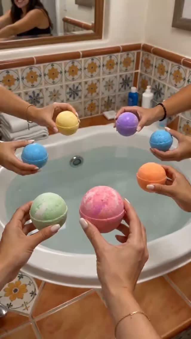Colorful spherical bath bombs held over a white bathtub in a tiled bathroom.
