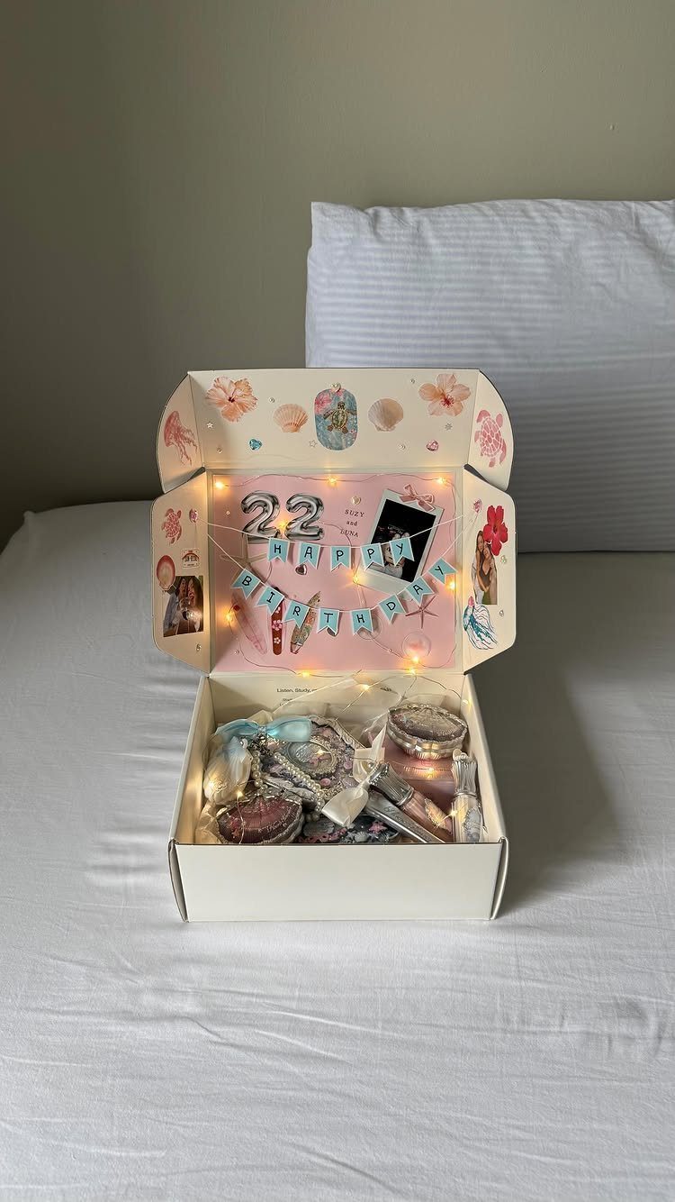 Open white gift box with skincare products and birthday decorations on a bed.