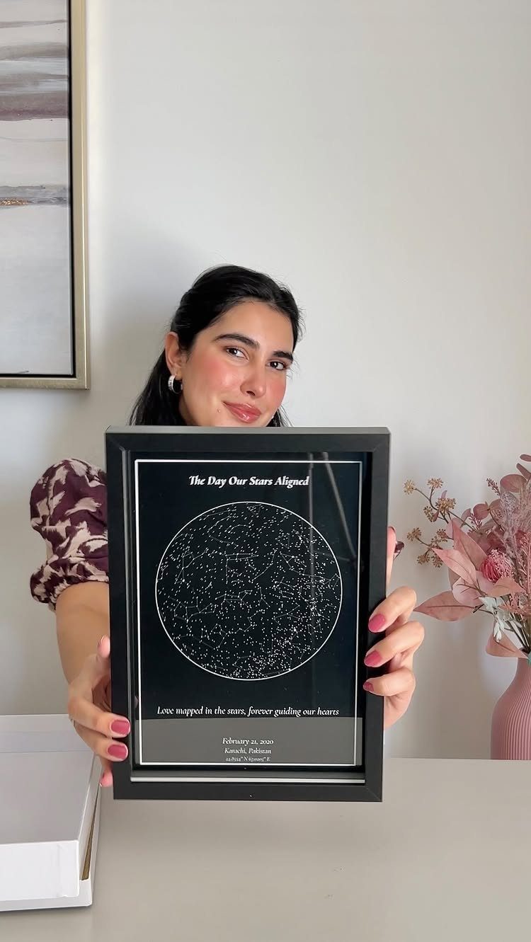 Woman holding a framed personalized star map canvas with white text on black.